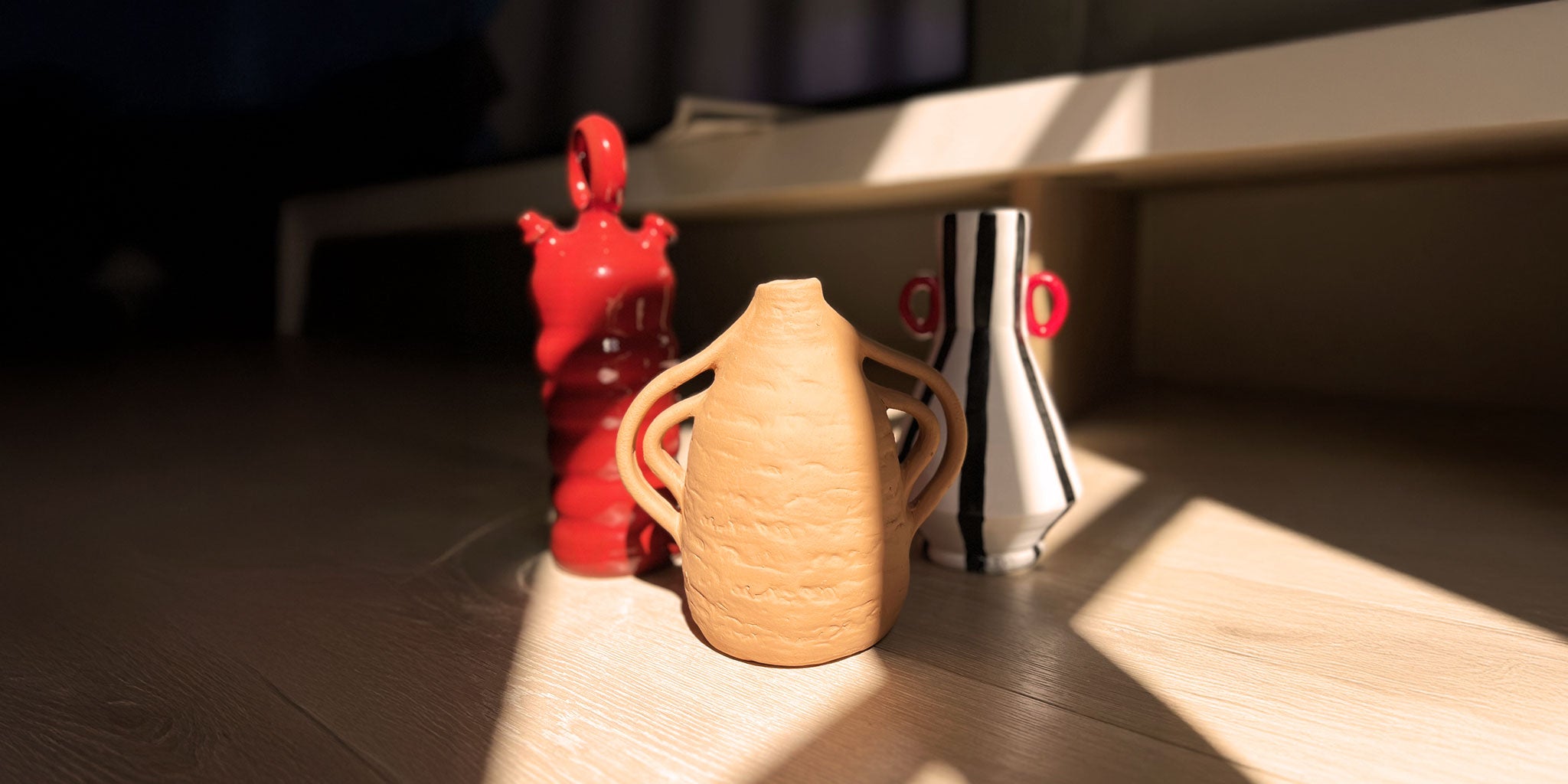 Trio di vasi artigianali in ceramica, uno in terracotta, uno rosso lucido e uno con strisce bianche e nere, illuminati da un raggio di sole su un pavimento in legno chiaro.