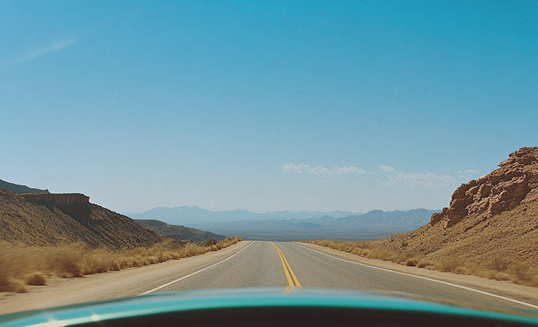 Vista di una strada deserta che si estende verso l'orizzonte, circondata da colline rocciose e paesaggi aridi, sotto un cielo azzurro limpido, catturata dal parabrezza di un'auto.